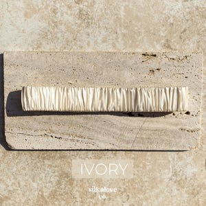 K&ouml;nnte beinhalten: Elfenbeinfarbenes, gerafftes Stoffaccessoire. Das rechteckige St&uuml;ck befindet sich auf einer strukturierten, hellfarbigen Steinoberfl&auml;che. Das Wort "IVORY" wird in wei&szlig;er, serifenloser Schrift angezeigt. Der Markenname "silknlove co." steht darunter.