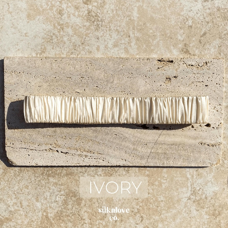 K&ouml;nnte beinhalten: Elfenbeinfarbenes, gerafftes Stoffaccessoire. Das rechteckige St&uuml;ck befindet sich auf einer strukturierten, hellfarbigen Steinoberfl&auml;che. Das Wort "IVORY" wird in wei&szlig;er, serifenloser Schrift angezeigt. Der Markenname "silknlove co." steht darunter.