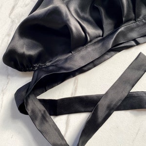 May include: A black satin bonnet with a tie closure. The bonnet is made of a shiny, smooth fabric and is designed to protect hair. The tie is long and can be secured around the head. The product is on a white marble surface.