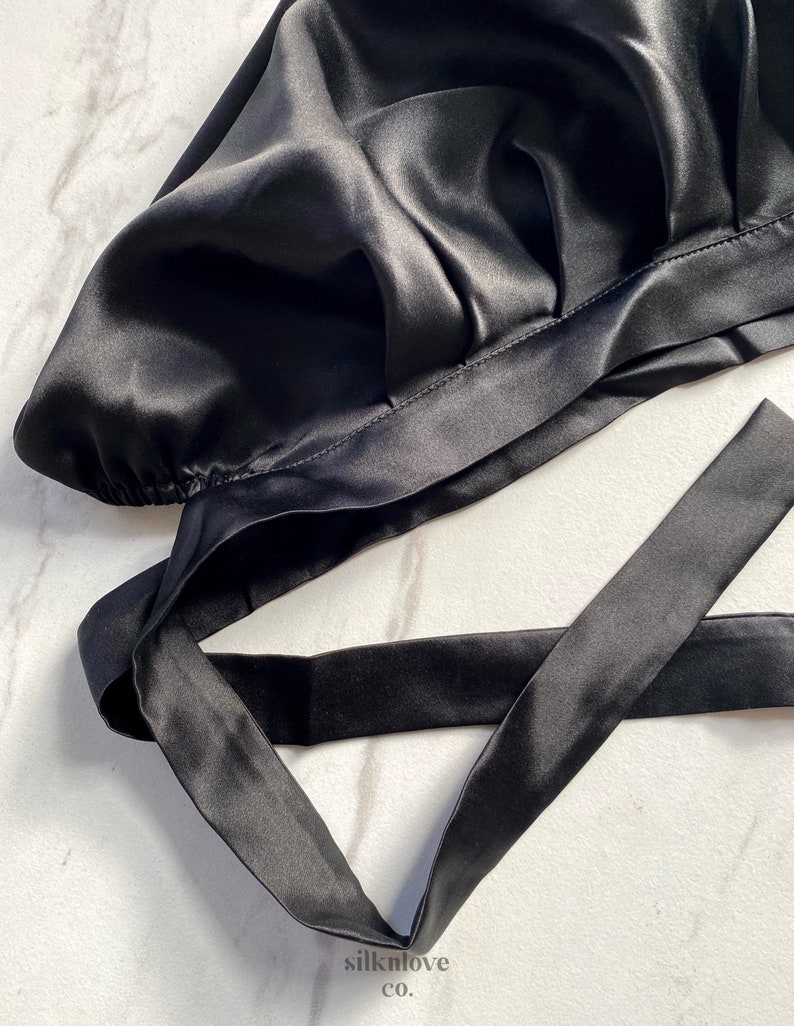 May include: A black satin bonnet with a tie closure. The bonnet is made of a shiny, smooth fabric and is designed to protect hair. The tie is long and can be secured around the head. The product is on a white marble surface.