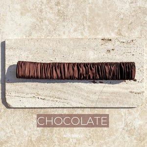 K&ouml;nnte beinhalten: Ein schokoladenfarbenes Haargummi aus seidigem Stoff. Das Haargummi ist gerafft und ger&uuml;scht, wodurch es eine strukturierte Optik erh&auml;lt. Das Wort "CHOCOLATE" ist in einem rechteckigen Feld darunter gedruckt.