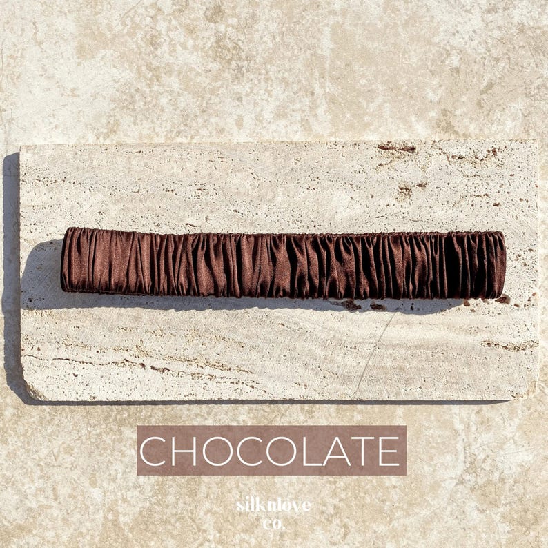K&ouml;nnte beinhalten: Ein schokoladenfarbenes Haargummi aus seidigem Stoff. Das Haargummi ist gerafft und ger&uuml;scht, wodurch es eine strukturierte Optik erh&auml;lt. Das Wort "CHOCOLATE" ist in einem rechteckigen Feld darunter gedruckt.