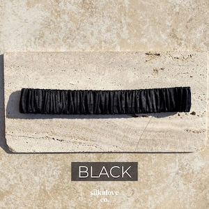 K&ouml;nnte beinhalten: Ein schwarzes, gerafftes Stoffaccessoire, m&ouml;glicherweise ein Stirnband oder ein Haargummi, liegt auf einer strukturierten, hellfarbigen Oberfl&auml;che. Das Wort "BLACK" wird in wei&szlig;er Schrift auf dunklem Hintergrund angezeigt. Der Markenname "silknlove co." steht darunter.