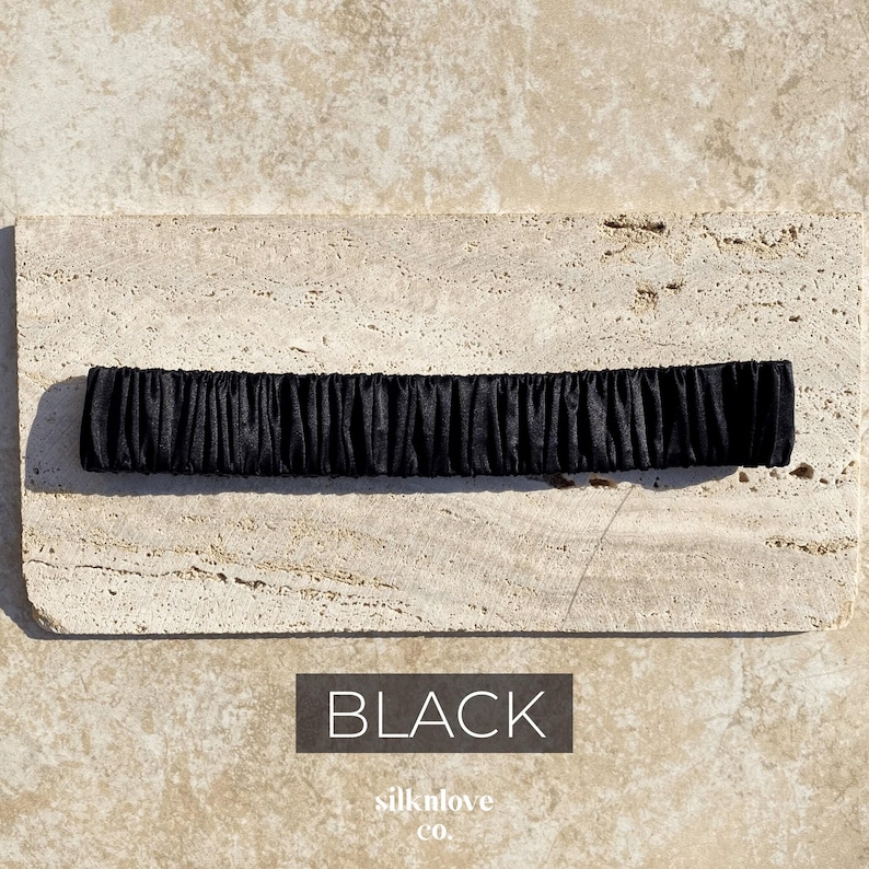 K&ouml;nnte beinhalten: Ein schwarzes, gerafftes Stoffaccessoire, m&ouml;glicherweise ein Stirnband oder ein Haargummi, liegt auf einer strukturierten, hellfarbigen Oberfl&auml;che. Das Wort "BLACK" wird in wei&szlig;er Schrift auf dunklem Hintergrund angezeigt. Der Markenname "silknlove co." steht darunter.