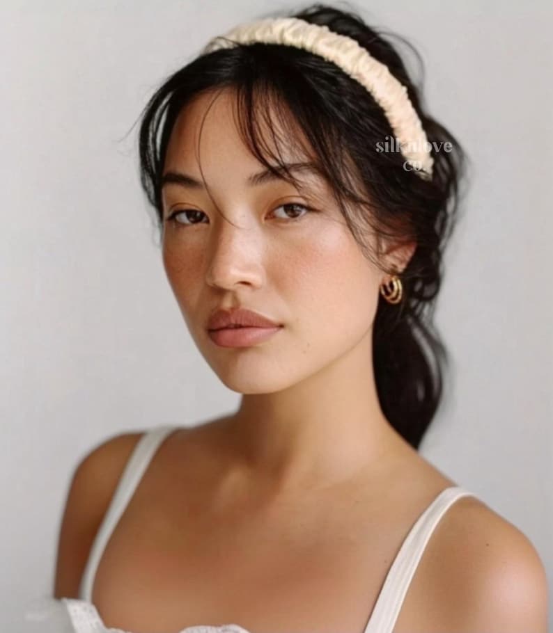 K&ouml;nnte beinhalten: Eine Frau mit dunklem Haar tr&auml;gt ein cremefarbenes Seidenstirnband. Das Stirnband ist breit und gerafft. Sie tr&auml;gt goldene Creolen und ein wei&szlig;es Tanktop. Der Text "silk love co." ist auf der rechten Seite des Bildes zu sehen.
