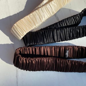 K&ouml;nnte beinhalten: Drei Seiden-Haarb&auml;nder in Creme, Schwarz und Braun. Die Haarb&auml;nder sind aus gl&auml;nzendem, gerafftem Stoff gefertigt. Das braune Haarband hat ein kleines Etikett mit der Aufschrift "silk move co."