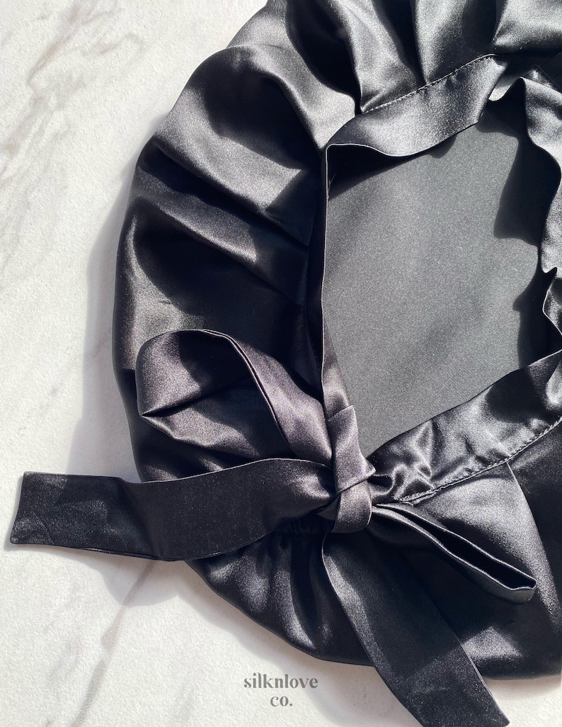 May include: A black satin bonnet with a tied bow closure. The bonnet is made of a shiny, smooth fabric and is designed to protect hair. The text "silknlove co." is visible in the lower right corner.