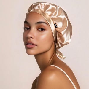 May include: A satin bonnet in a light gold color. The bonnet has a rounded crown and a satin tie at the nape of the neck. A small black tag is visible on the side of the bonnet. The bonnet is designed to protect hair.