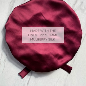 May include: A burgundy satin sleep cap with a tie. The cap is made with the finest 22 momme mulberry silk. The text "MADE WITH THE FINEST 22 MOMME MULBERRY SILK" is printed on a white rectangle in the center of the cap. The text "silklove co." is printed below the cap.