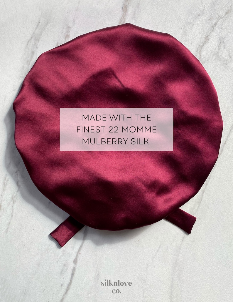 May include: A burgundy satin sleep cap with a tie. The cap is made with the finest 22 momme mulberry silk. The text "MADE WITH THE FINEST 22 MOMME MULBERRY SILK" is printed on a white rectangle in the center of the cap. The text "silklove co." is printed below the cap.