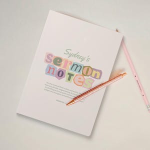 May include: A white notebook with the text "Sydney's sermon notes" in colorful, cut-out style letters. A rose gold glitter pen and a pink pencil with the text "Radiate Positivity" are next to the notebook. The notebook has a quote from Hebrews 4:12.