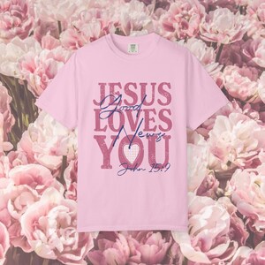 Puede incluir: Una camiseta rosa claro con el texto "JESUS LOVES YOU" en un diseño desgastado y en capas. La camiseta también incluye el versículo bíblico Juan 15:9. El fondo presenta flores rosas.