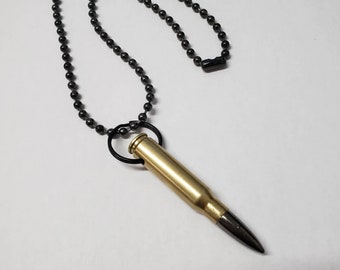 Può includere: Una collana con un ciondolo a forma di bossolo di proiettile dorato e una catena nera. Il bossolo di proiettile è attaccato alla catena con un anello di metallo nero.
