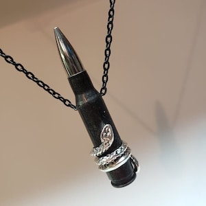 May include: A black bullet casing necklace with a silver snake charm wrapped around it. The necklace is on a black chain.