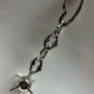 May include: A silver-toned metal keychain featuring a polished spherical charm with six protruding spikes attached to a short, thick chain. The chain consists of three irregular, organic-shaped links connected to a larger, elongated carabiner clasp.