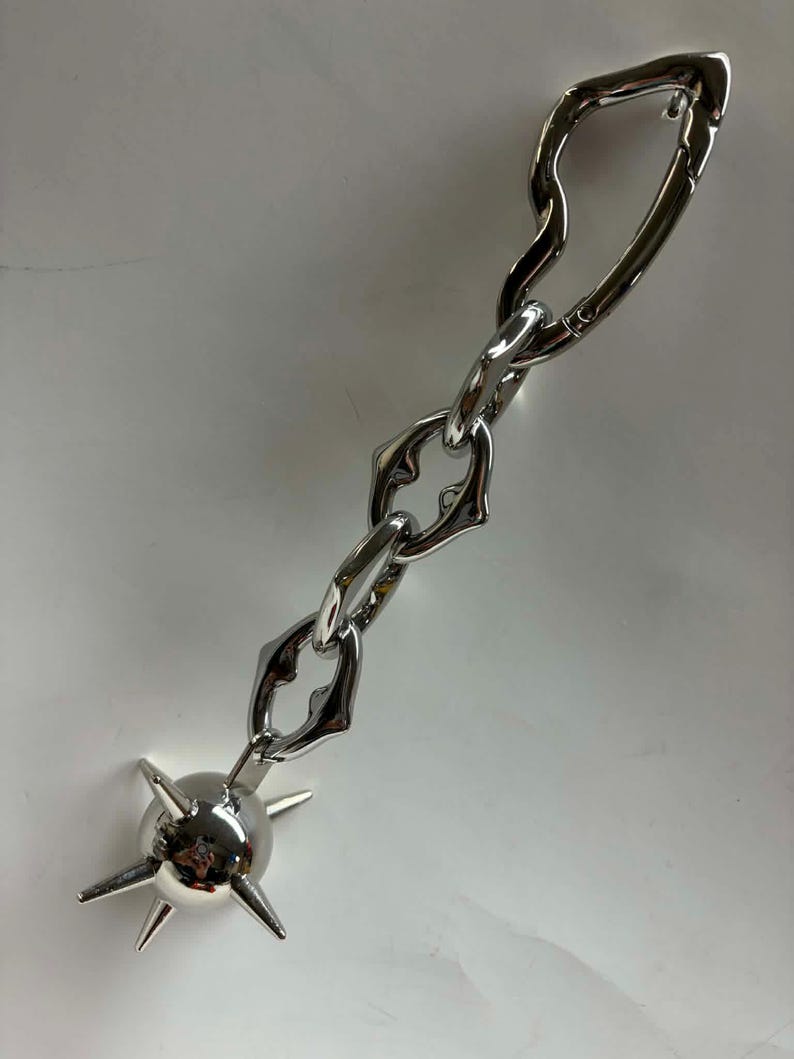 May include: A silver-toned metal keychain featuring a polished spherical charm with six protruding spikes attached to a short, thick chain. The chain consists of three irregular, organic-shaped links connected to a larger, elongated carabiner clasp.