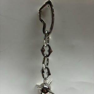 May include: A silver-toned metal keychain featuring an irregular, organic-shaped top link connected to a short chain of three textured, angular links. The chain terminates in a spherical charm adorned with four protruding, pointed spikes.
