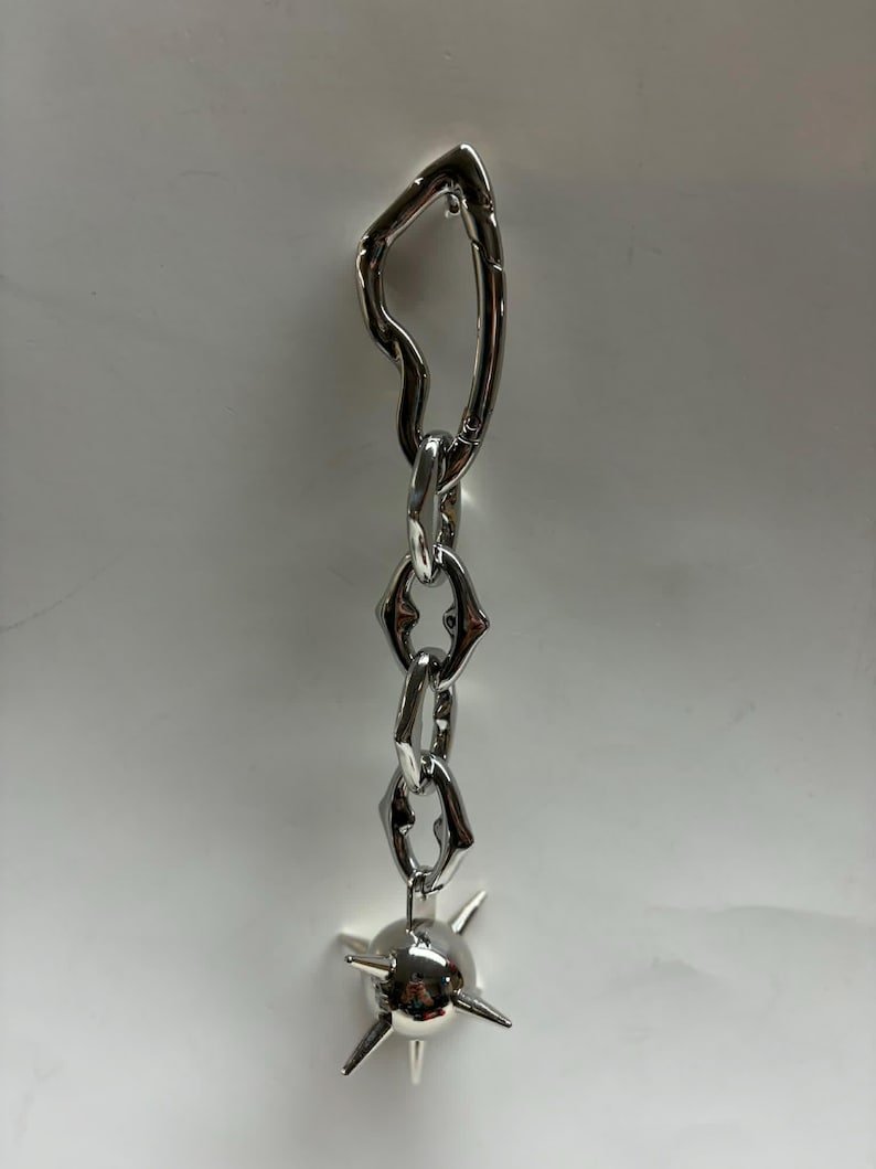 May include: A silver-toned metal keychain featuring an irregular, organic-shaped top link connected to a short chain of three textured, angular links. The chain terminates in a spherical charm adorned with four protruding, pointed spikes.