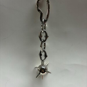 May include: A silver-toned metal keychain featuring an irregular, organic-shaped carabiner clip attached to a short chain of three stylized, angular links. The chain terminates in a spherical charm adorned with several protruding, sharp spikes.
