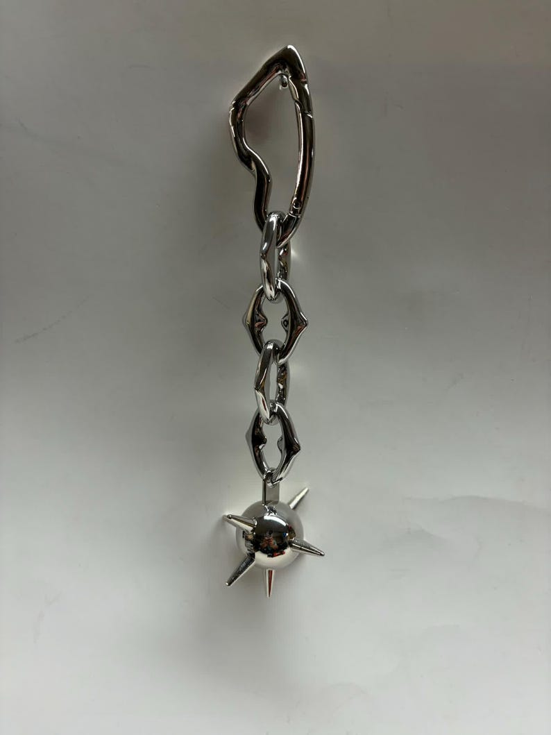 May include: A silver-toned metal keychain featuring an irregular, organic-shaped carabiner clip attached to a short chain of three stylized, angular links. The chain terminates in a spherical charm adorned with several protruding, sharp spikes.