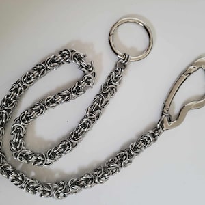 K&ouml;nnte beinhalten: Eine silberfarbene Kette mit einem gro&szlig;en Ring und einem Verschluss. Die Kette besteht aus ineinandergreifenden Metallringen, wodurch ein strukturiertes, gewebtes Aussehen entsteht. Ring und Verschluss sind ebenfalls silberfarben, vor wei&szlig;em Hintergrund.