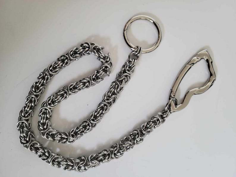 K&ouml;nnte beinhalten: Eine silberfarbene Kette mit einem gro&szlig;en Ring und einem Verschluss. Die Kette besteht aus ineinandergreifenden Metallringen, wodurch ein strukturiertes, gewebtes Aussehen entsteht. Ring und Verschluss sind ebenfalls silberfarben, vor wei&szlig;em Hintergrund.