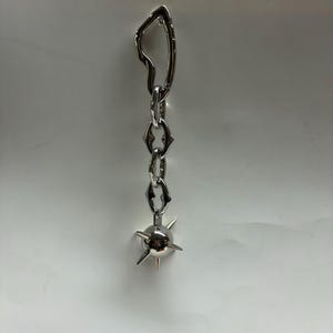 May include: A silver-toned metal earring featuring an irregular, open-shaped top link connected to a short chain of three pointed, diamond-shaped links. The chain terminates in a small, spherical charm adorned with several protruding, sharp spikes.