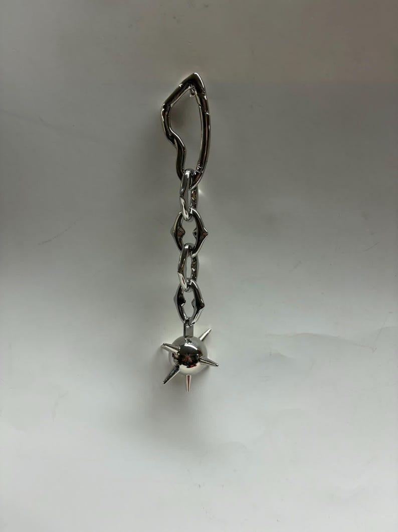 May include: A silver-toned metal earring featuring an irregular, open-shaped top link connected to a short chain of three pointed, diamond-shaped links. The chain terminates in a small, spherical charm adorned with several protruding, sharp spikes.