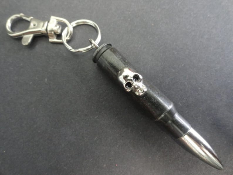 May include: A black bullet casing keychain with a silver skull design. The keychain has a silver split ring and a silver lobster clasp.