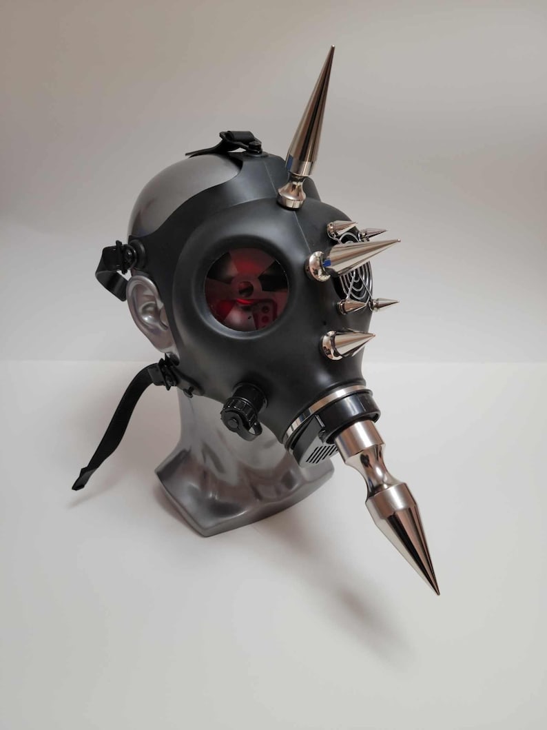 May include: A black gas mask with silver accents and multiple silver spikes. The mask has a clear lens with a red interior and is mounted on a gray mannequin head. The mask's design is industrial and edgy.