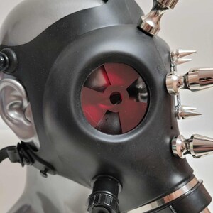 May include: Black gas mask with silver accents and metallic spikes. The mask features a red, fan-shaped filter and adjustable straps. The mask is displayed on a mannequin head.