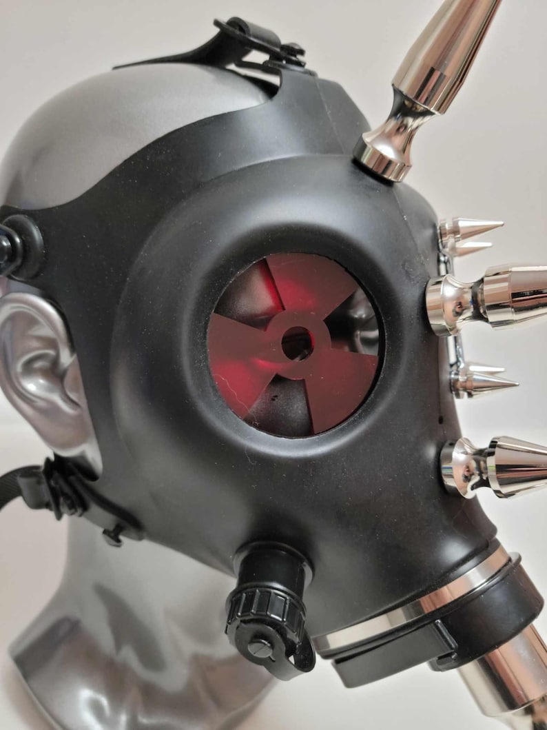 May include: Black gas mask with silver accents and metallic spikes. The mask features a red, fan-shaped filter and adjustable straps. The mask is displayed on a mannequin head.