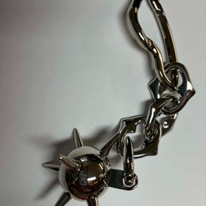 May include: A silver-toned metal keychain featuring a spherical mace charm with multiple pointed spikes attached to a chain of irregular, organic-shaped links and a large, elongated carabiner clip with a spring-loaded gate mechanism.