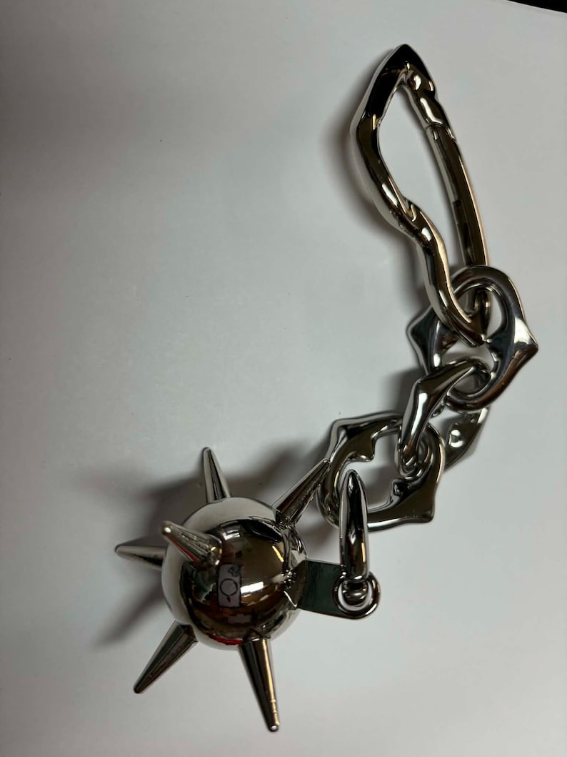 May include: A silver-toned metal keychain featuring a spherical mace charm with multiple pointed spikes attached to a chain of irregular, organic-shaped links and a large, elongated carabiner clip with a spring-loaded gate mechanism.