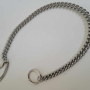 May include: A silver-colored chain with a heart-shaped clasp and a circular clasp. The chain is made of interlocking links and is displayed on a white surface. The chain is a jewelry accessory.