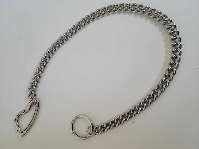 May include: A silver-colored chain with a heart-shaped clasp and a circular clasp. The chain is made of interlocking links and is displayed on a white surface. The chain is a jewelry accessory.