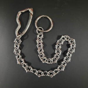 May include: A silver wallet chain featuring a carabiner clip and a key ring. The chain is composed of interlocking links with a star-shaped pattern. The carabiner clip has a curved design.