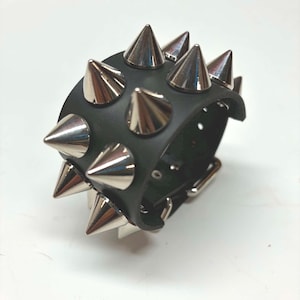 Könnte beinhalten: Ein schwarzes Armband mit zwei Reihen großer, polierter, silberfarbener Metallspitzen. Das Band besteht aus einem glatten, matten Material und verfügt über einen seitlich sichtbaren Metall-Schnallenverschluss für einen sicheren Halt.