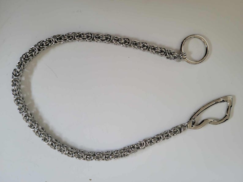 K&ouml;nnte beinhalten: Silberfarbene Kette mit einem kreisf&ouml;rmigen Ring an einem Ende und einem gebogenen Verschluss am anderen. Die Kette besteht aus kleinen, miteinander verbundenen Gliedern, die ein strukturiertes Aussehen erzeugen. Es ist wahrscheinlich ein Schmuckbestandteil oder Zubeh&ouml;r.