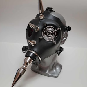 May include: Black gas mask with silver spikes and accents. The mask features a silver fan cover, a silver beak-like attachment, and a large spike on top. The mask is displayed on a gray mannequin head.