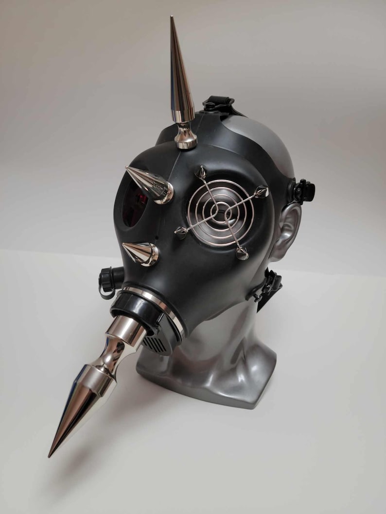 May include: Black gas mask with silver spikes and accents. The mask features a silver fan cover, a silver beak-like attachment, and a large spike on top. The mask is displayed on a gray mannequin head.