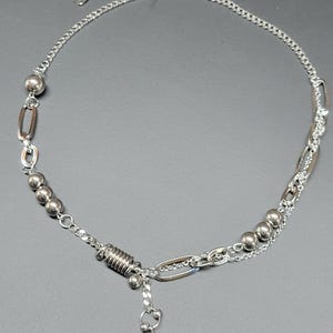 May include: Silver necklace with a mix of chain styles and decorative elements. The necklace features small beads, oval links, and a dangling conical pendant. The design includes a spring-like coil and a delicate chain. The necklace is made of silver.