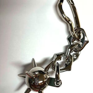 May include: A silver-toned metal keychain featuring a spherical charm with multiple protruding spikes attached to a short, thick, irregular chain link segment that connects to a large, elongated, asymmetrical carabiner-style clip.