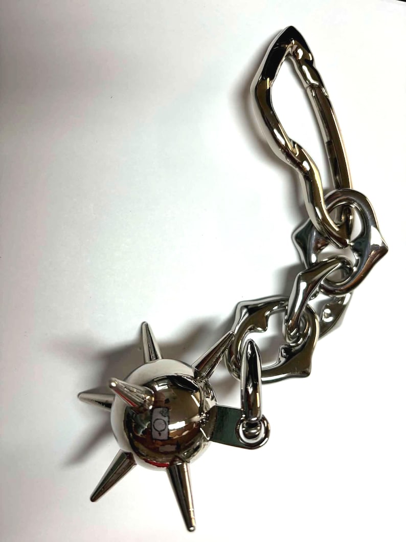 May include: A silver-toned metal keychain featuring a spherical charm with multiple protruding spikes attached to a short, thick, irregular chain link segment that connects to a large, elongated, asymmetrical carabiner-style clip.