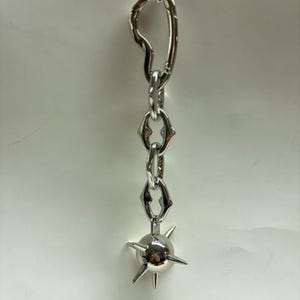 May include: A silver-toned metal keychain featuring an irregular heart-shaped carabiner at the top, connected to a short chain of three stylized links, and ending with a spherical charm adorned with several sharp, protruding metal spikes.