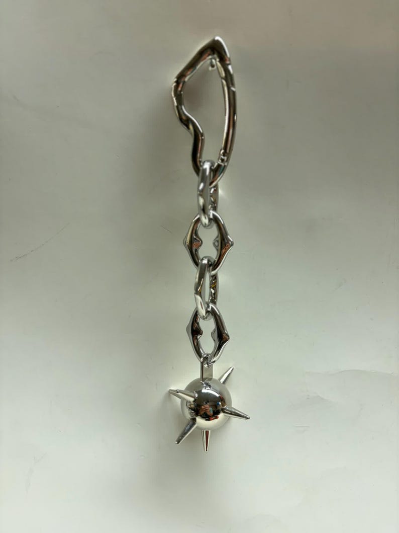 May include: A silver-toned metal keychain featuring an irregular heart-shaped carabiner at the top, connected to a short chain of three stylized links, and ending with a spherical charm adorned with several sharp, protruding metal spikes.