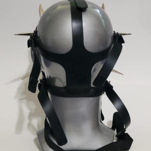 May include: A silver-colored mask with black straps and metal spikes. The mask has a rounded shape with a black strap across the top and around the face. The spikes are evenly spaced around the mask's perimeter.