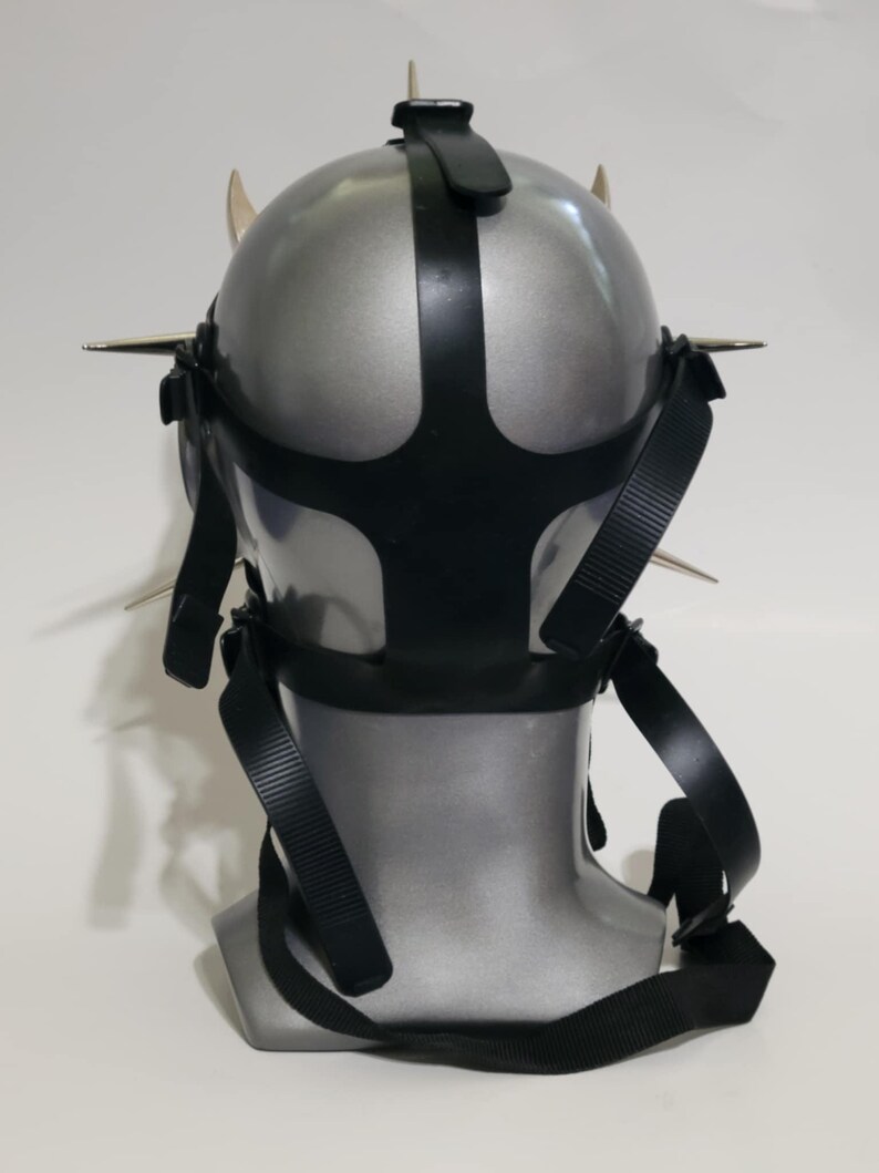 May include: A silver-colored mask with black straps and metal spikes. The mask has a rounded shape with a black strap across the top and around the face. The spikes are evenly spaced around the mask's perimeter.