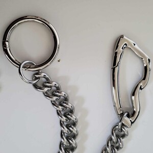 May include: Close-up of a silver metal chain with a large circular clasp and a curved carabiner-style clasp. The chain links are thick and interlocked, creating a sturdy appearance. The background is a plain white surface.