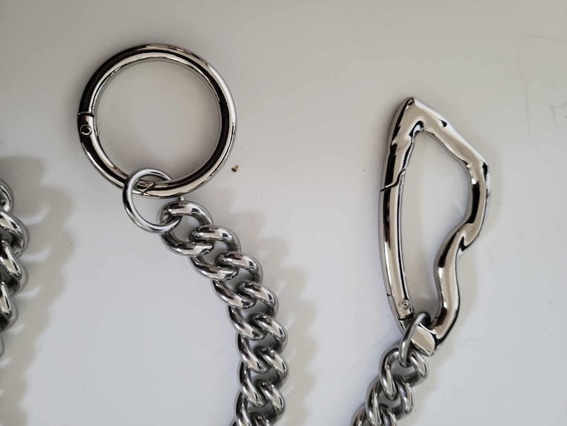 May include: Close-up of a silver metal chain with a large circular clasp and a curved carabiner-style clasp. The chain links are thick and interlocked, creating a sturdy appearance. The background is a plain white surface.